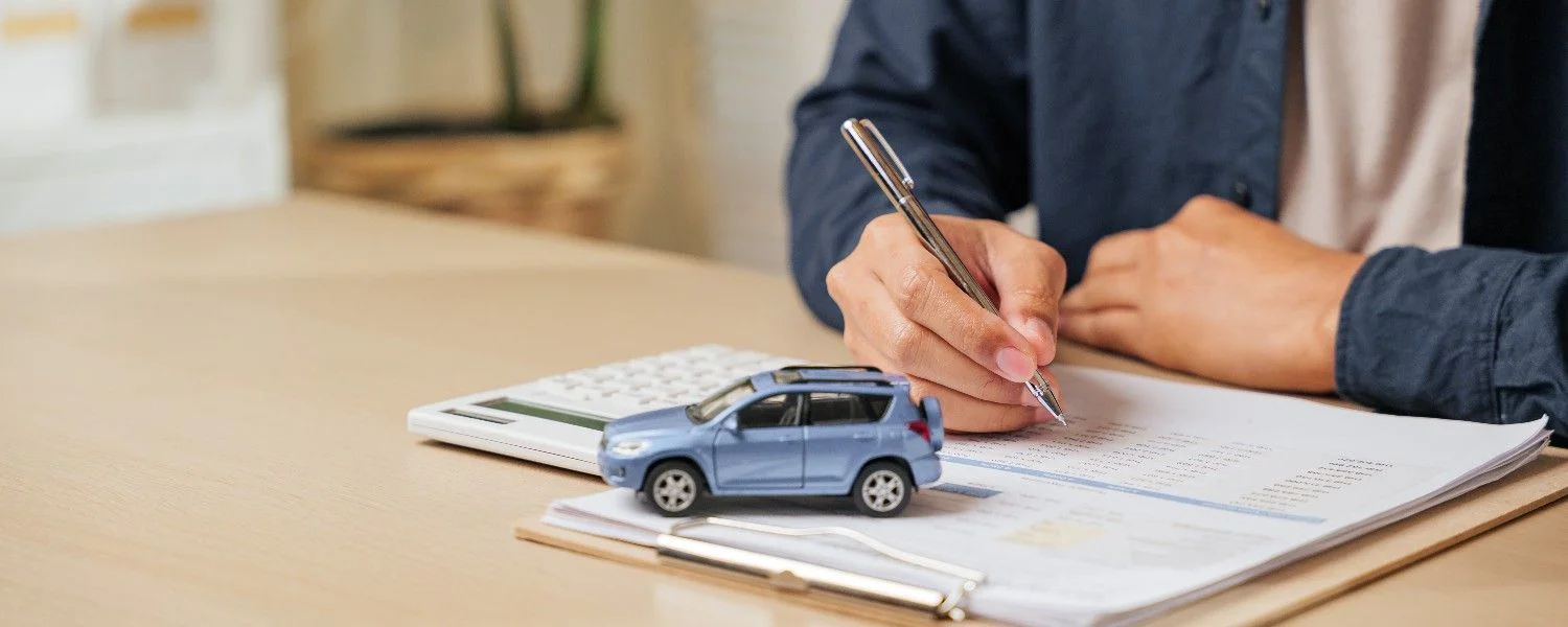 Person filling in paperwork at a desk beside a small model car